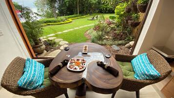 A dining table with two wicker chairs, a view of a garden with a lawn and trees, and a stone pathway.