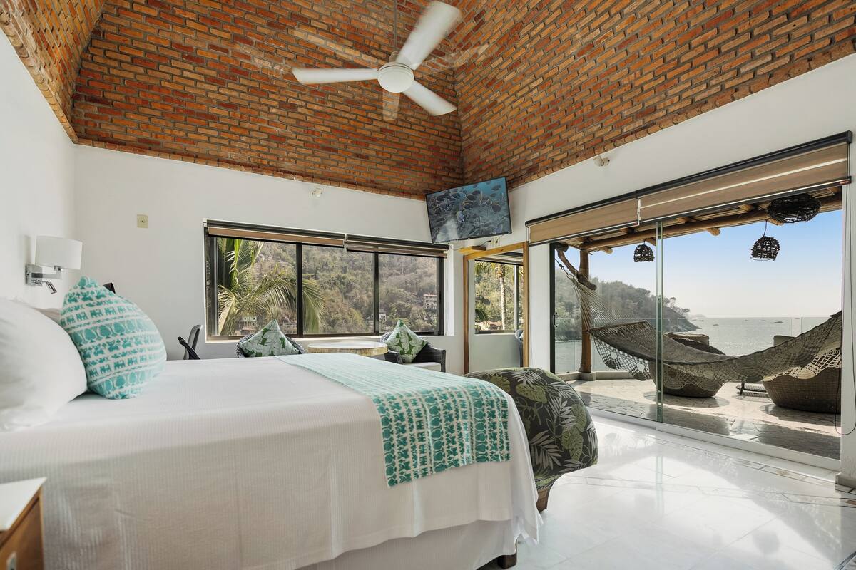 Un dormitorio con una cama grande, ventilador de techo, vista al mar y una hamaca en el exterior.