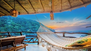 A hammock, wooden bench, and hanging light fixture in a room with a view of a forest and water.