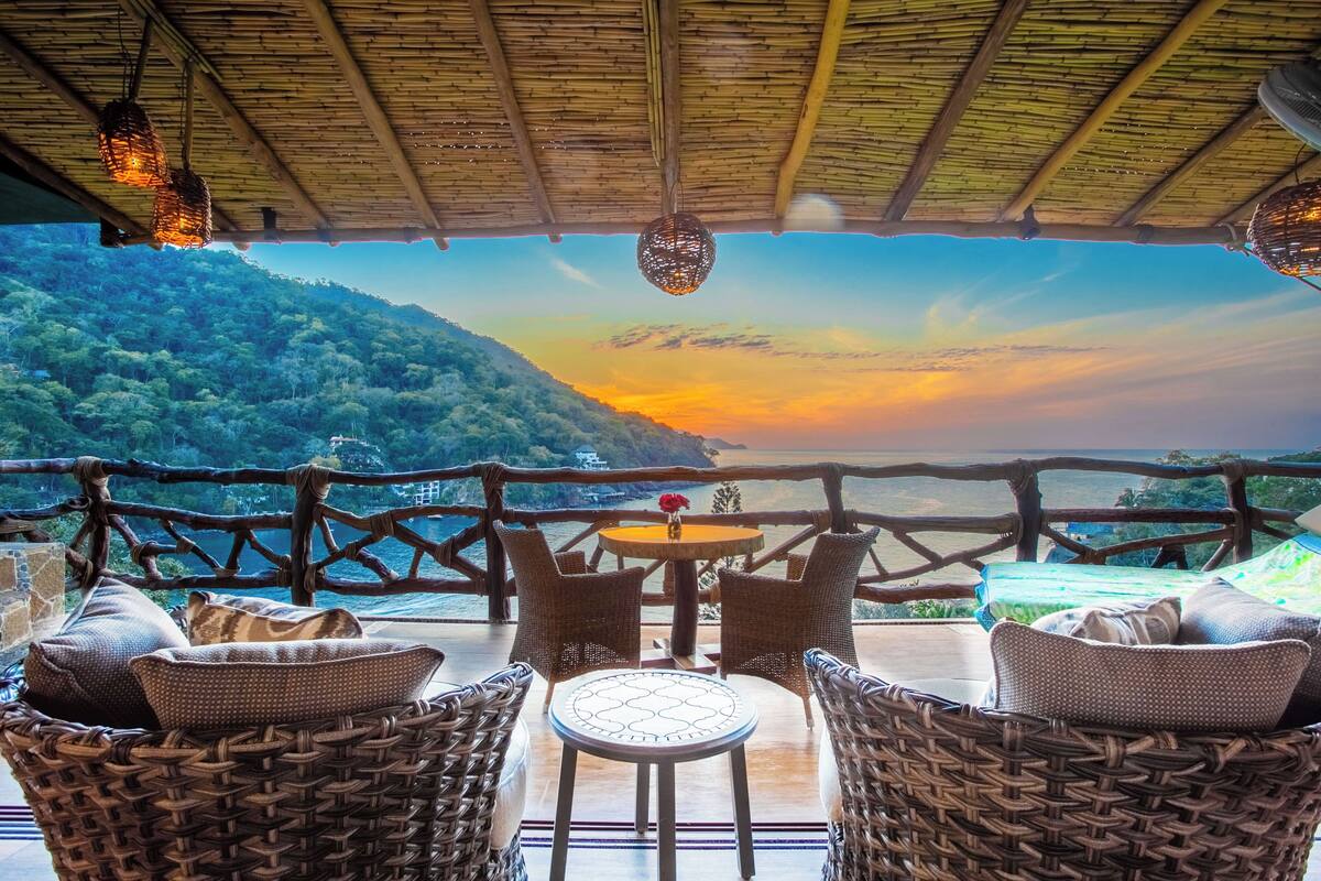 Un rincón de descanso de mimbre con una mesa y sillas, con vistas panorámicas a una ladera boscosa y una masa de agua al atardecer.