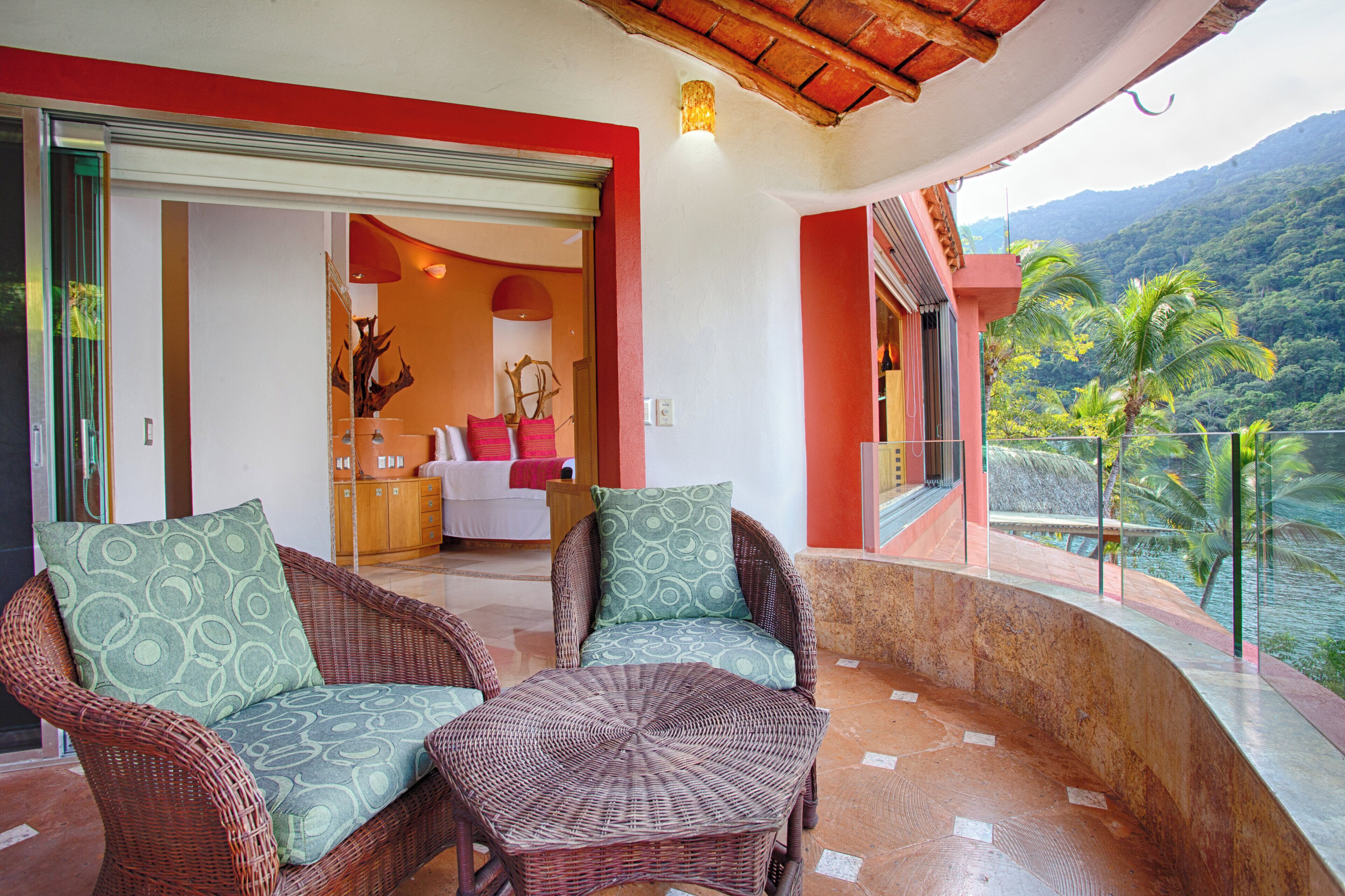 A balcony with wicker furniture, a view of a mountainous landscape, and a door leading to a room with a bed and a decorative vase.