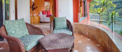 A balcony with wicker furniture, a view of a mountainous landscape, and a door leading to a room with a bed and a decorative vase.