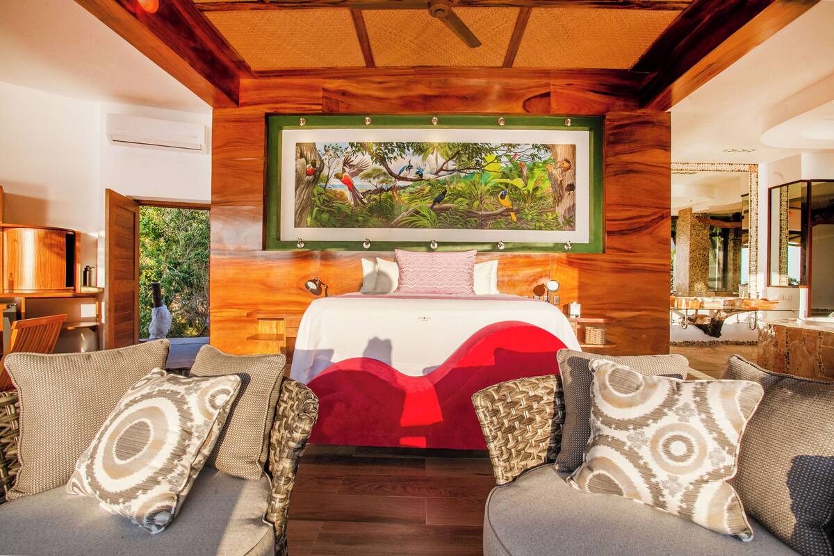 Un dormitorio con una cama grande, un cuadro de una escena tropical y vistas al exterior.
