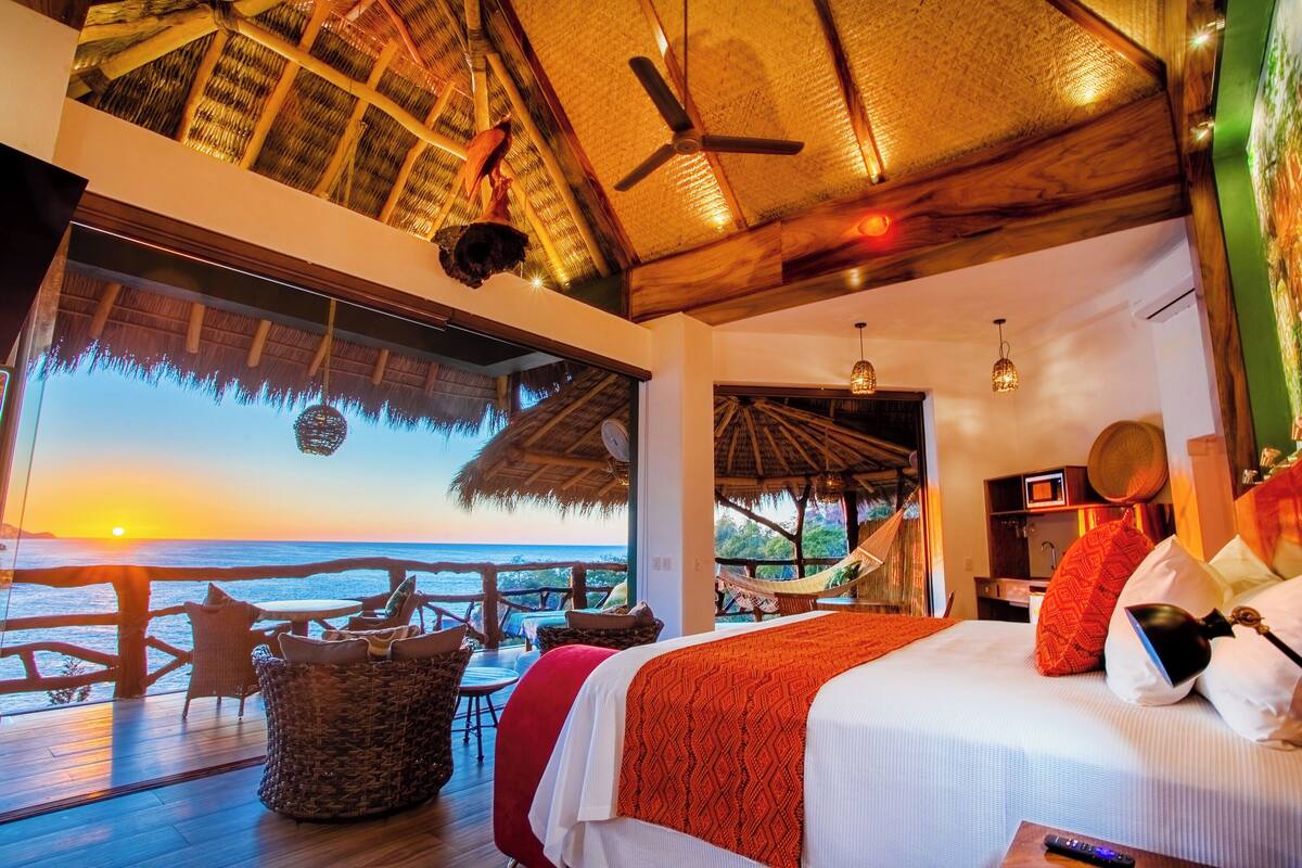 Una habitación con una cama, vistas al atardecer, techo de madera y una hamaca.