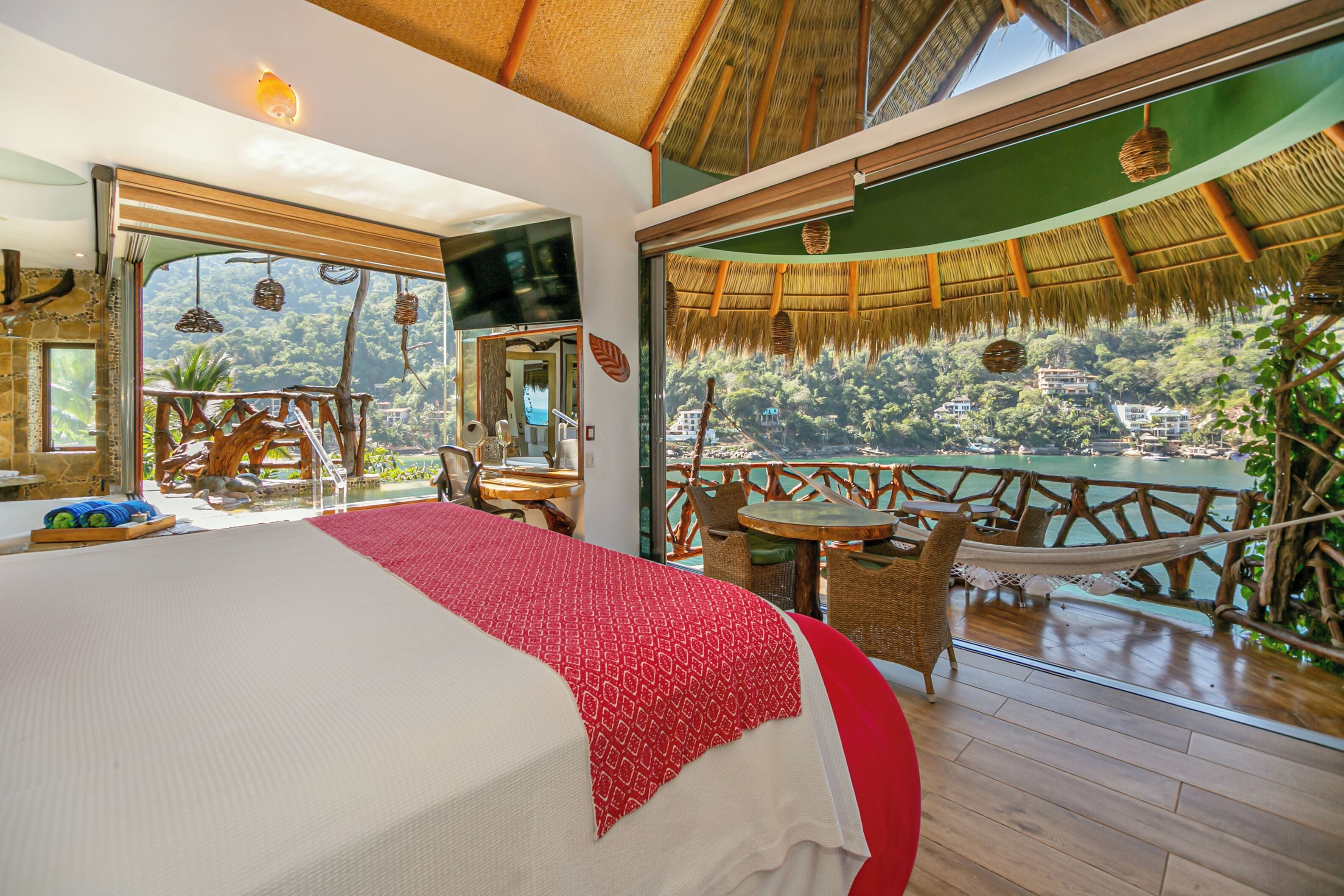 Una habitación con cama, televisión, balcón con mobiliario de mimbre y vistas a una zona boscosa.