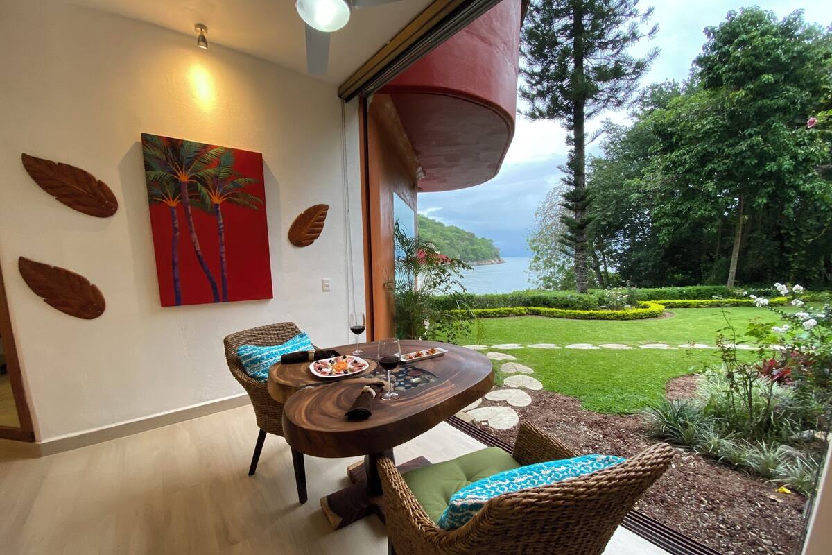 Un comedor con una mesa redonda de madera, sillas de mimbre y vistas a un jardín ajardinado con un cuerpo de agua.