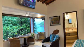 Een kamer met televisie, rieten meubels, een groot raam met uitzicht op groen en water, en een badkamer met wastafel en spiegel.