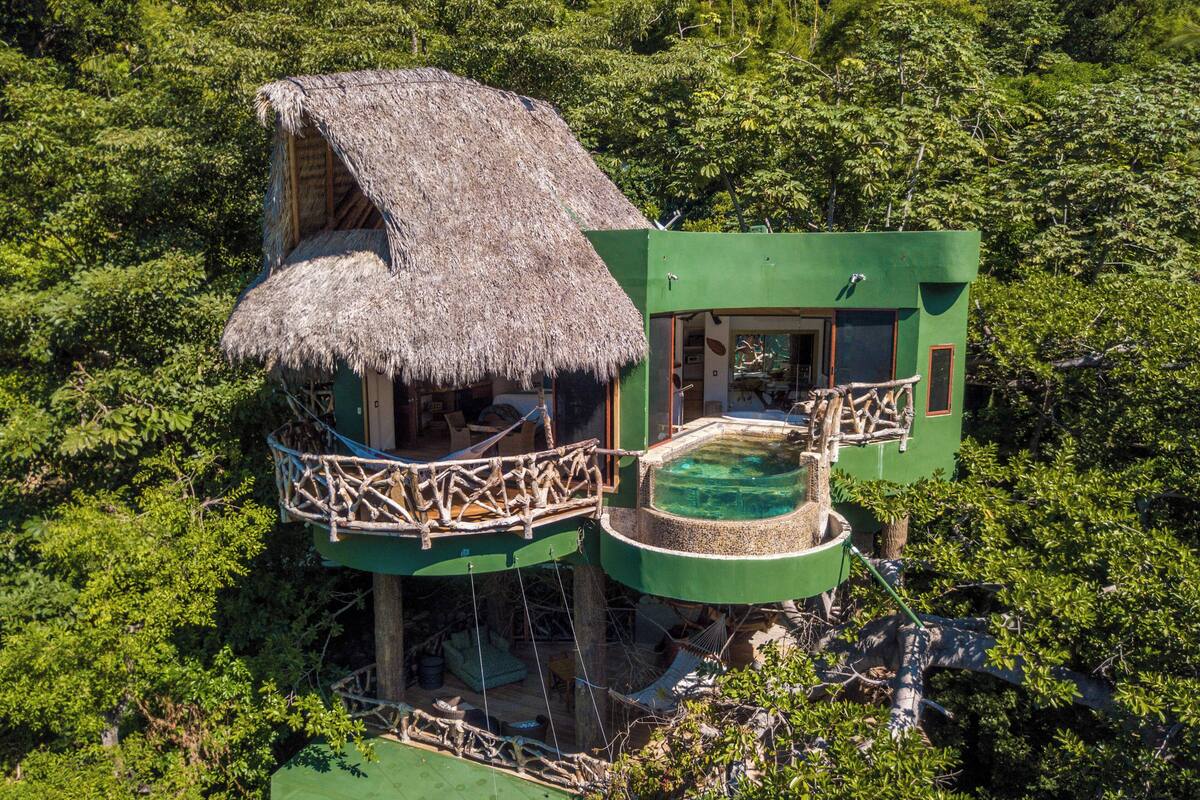 Una casa en árbol con techo de paja, piscina y balcón.