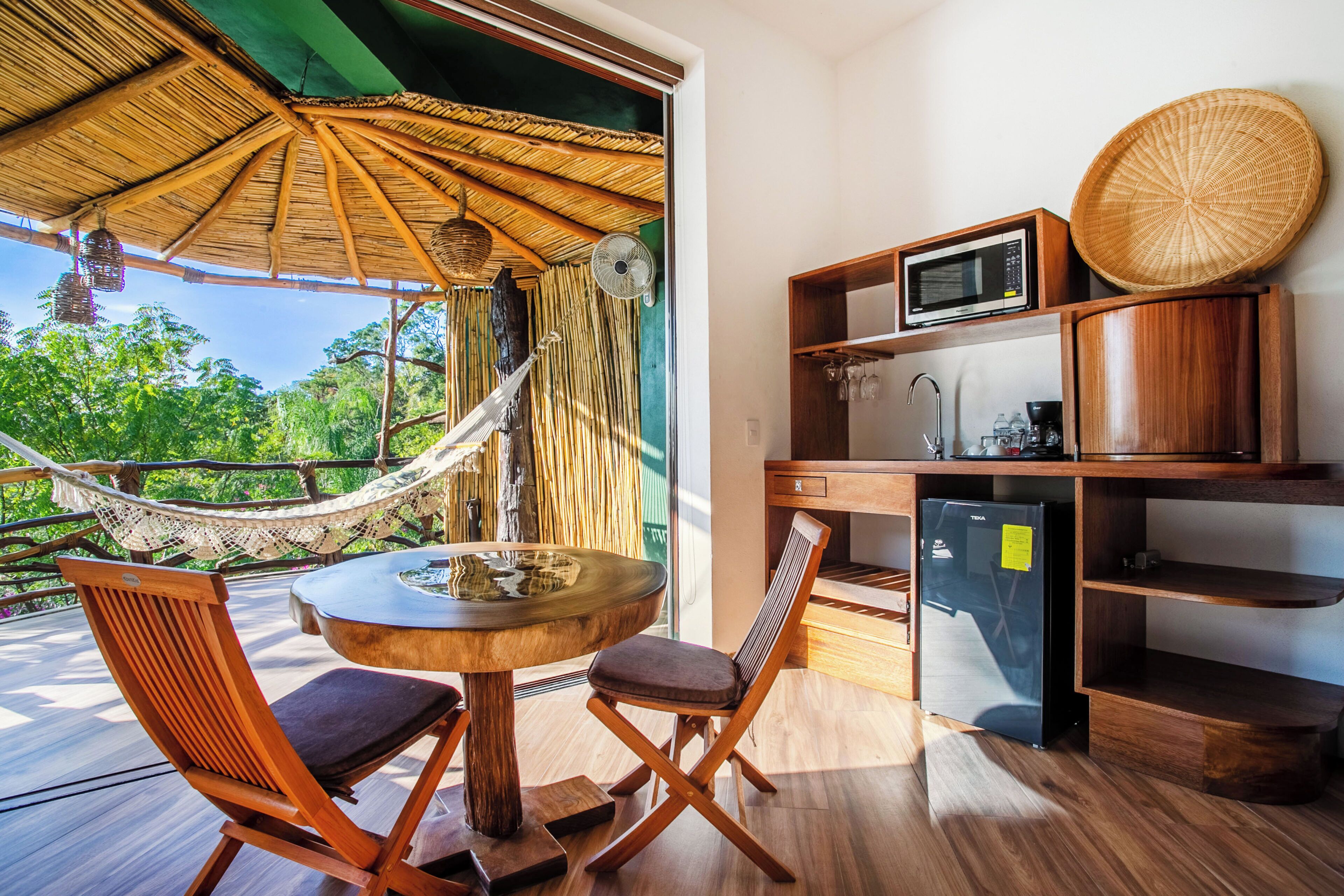 A room with a wooden dining table, two chairs, a hammock, a kitchen area with a microwave, and a refrigerator.