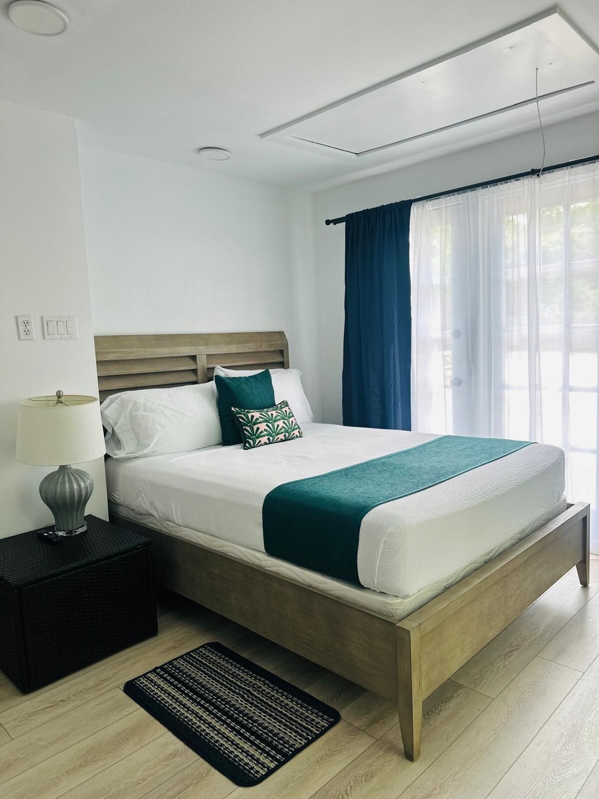 A modern bedroom with a wooden bed, white bedding, a green throw, a nightstand with a lamp, and a window with blue curtains.
