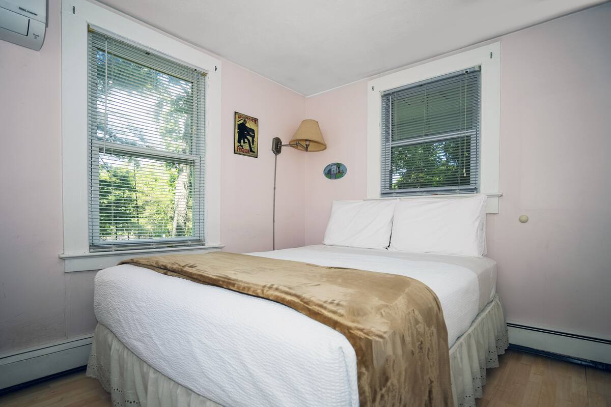 A bedroom with a bed, two windows with blinds, a floor lamp, and a wall-mounted air conditioner.