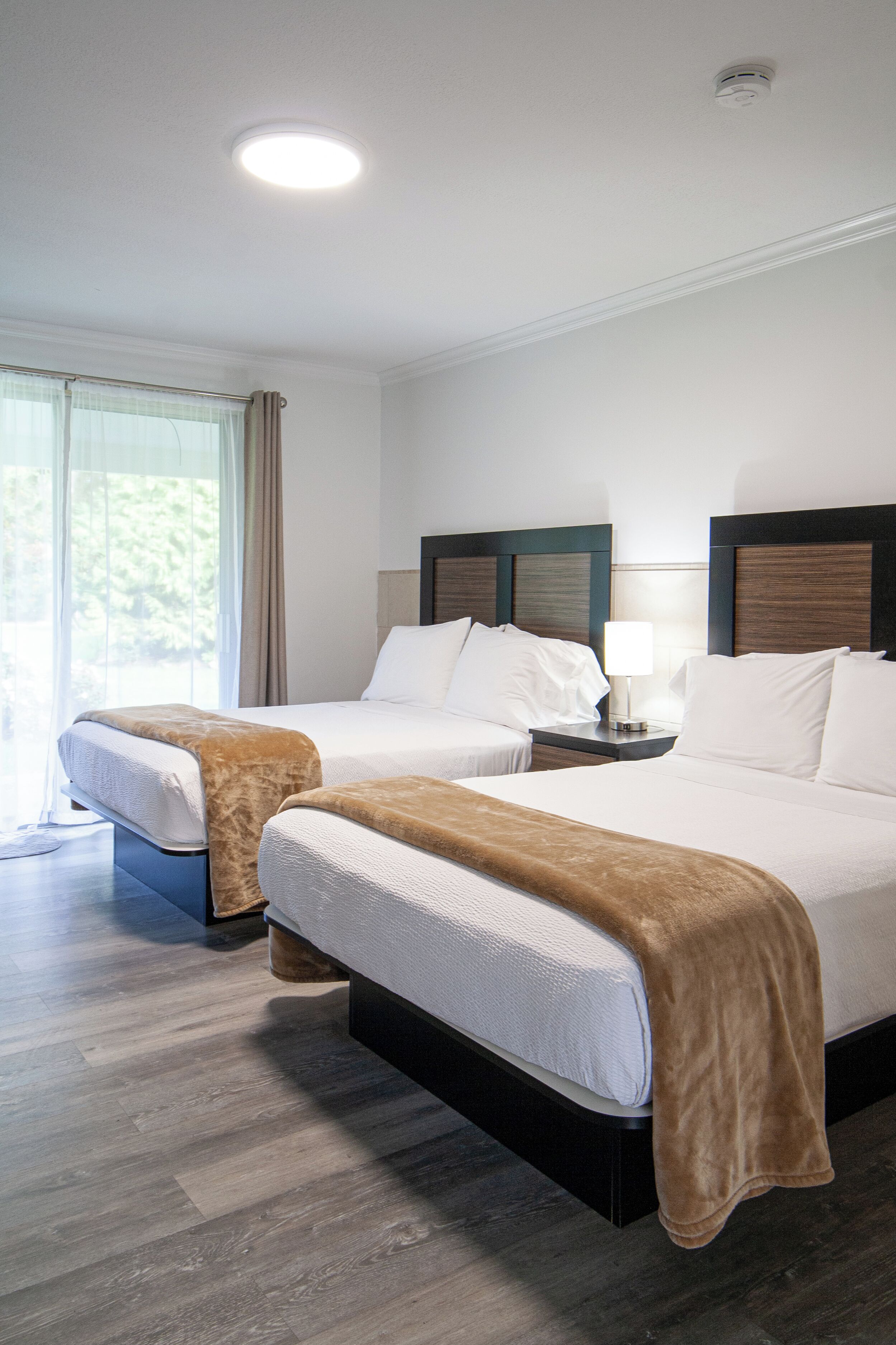 A hotel room with two beds, a large window with curtains, a ceiling light, and a wooden floor.