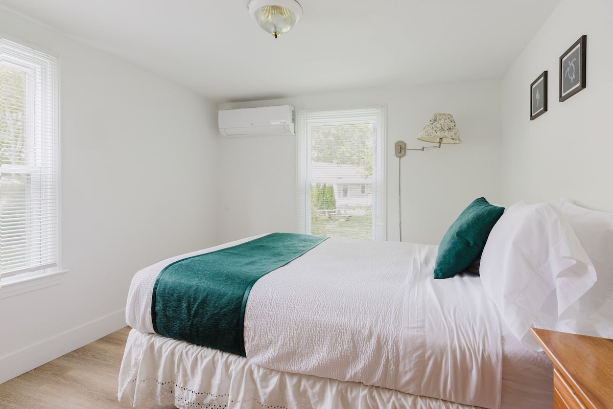 A bedroom with a bed, white walls, a window with blinds, an air conditioning unit, and a lamp.