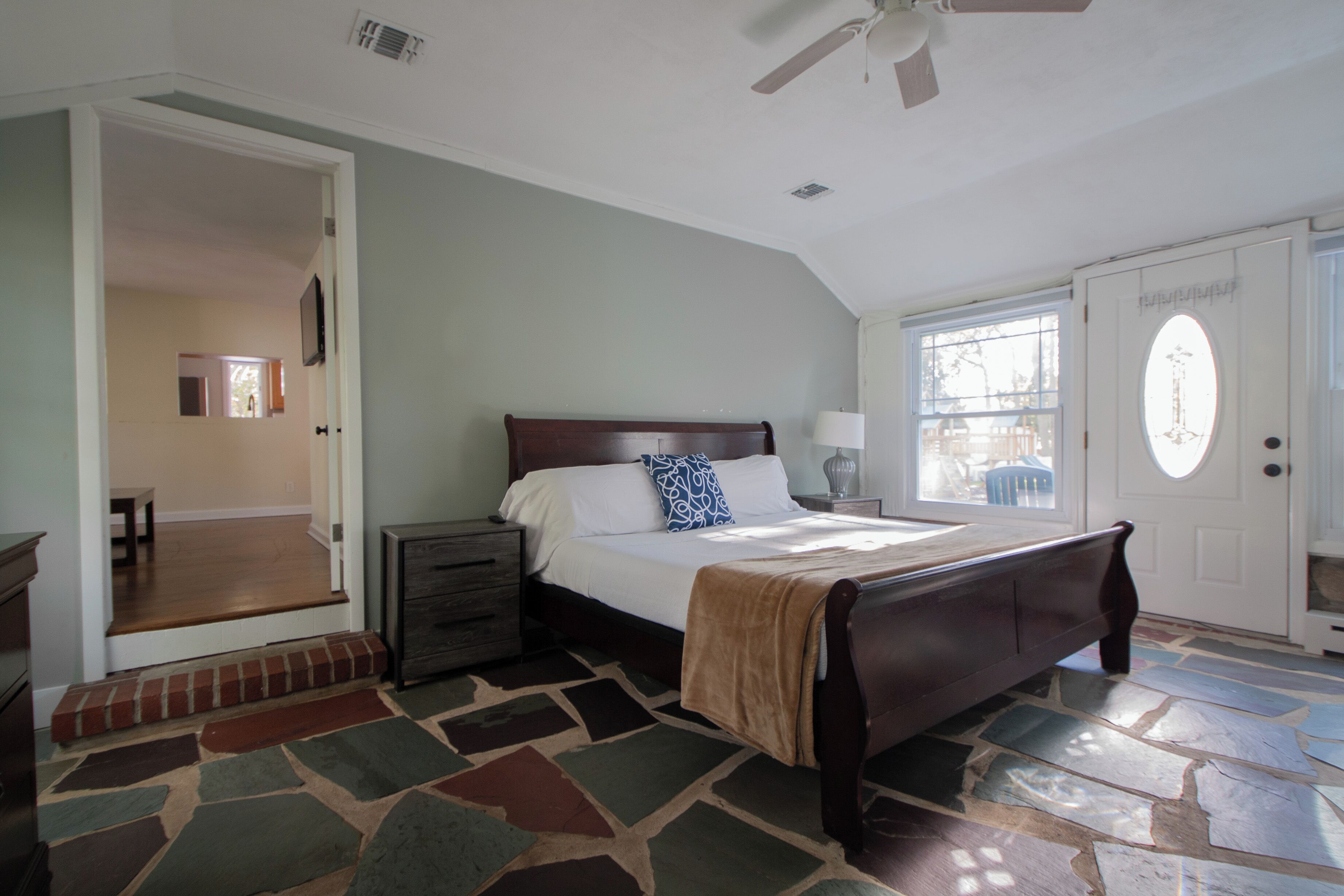 A bedroom with a large bed, a nightstand, a dresser, a door with a window, and a patterned rug.