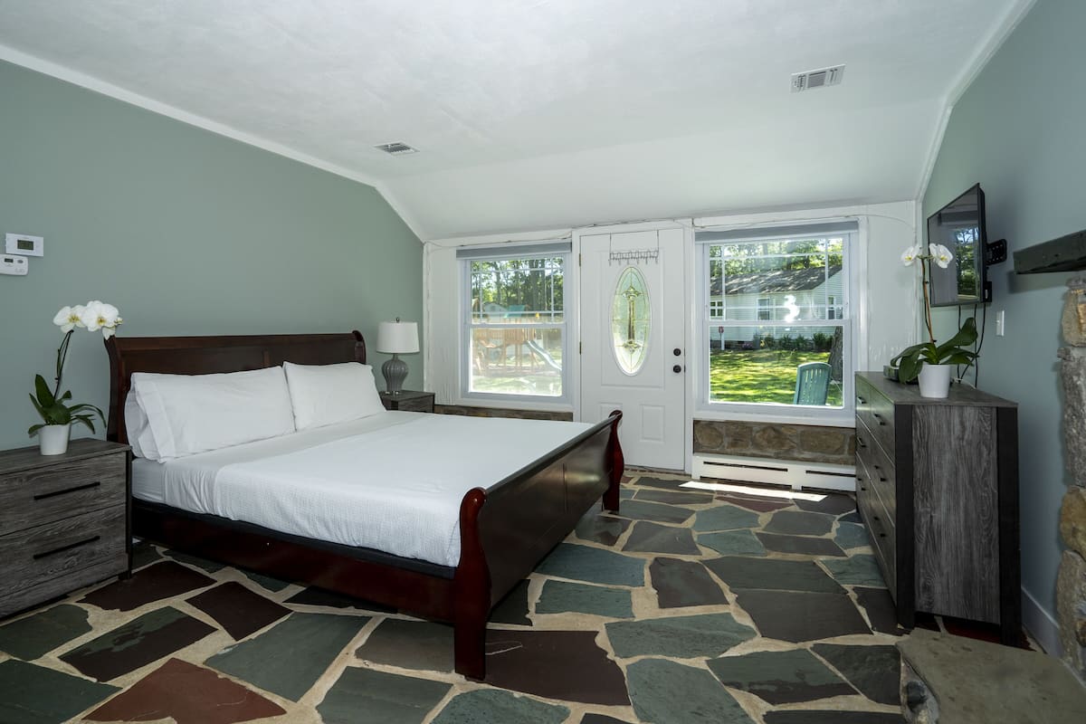 A bedroom with a stone-patterned floor, a wooden bed, a nightstand, a dresser, and a window with a view of a backyard.