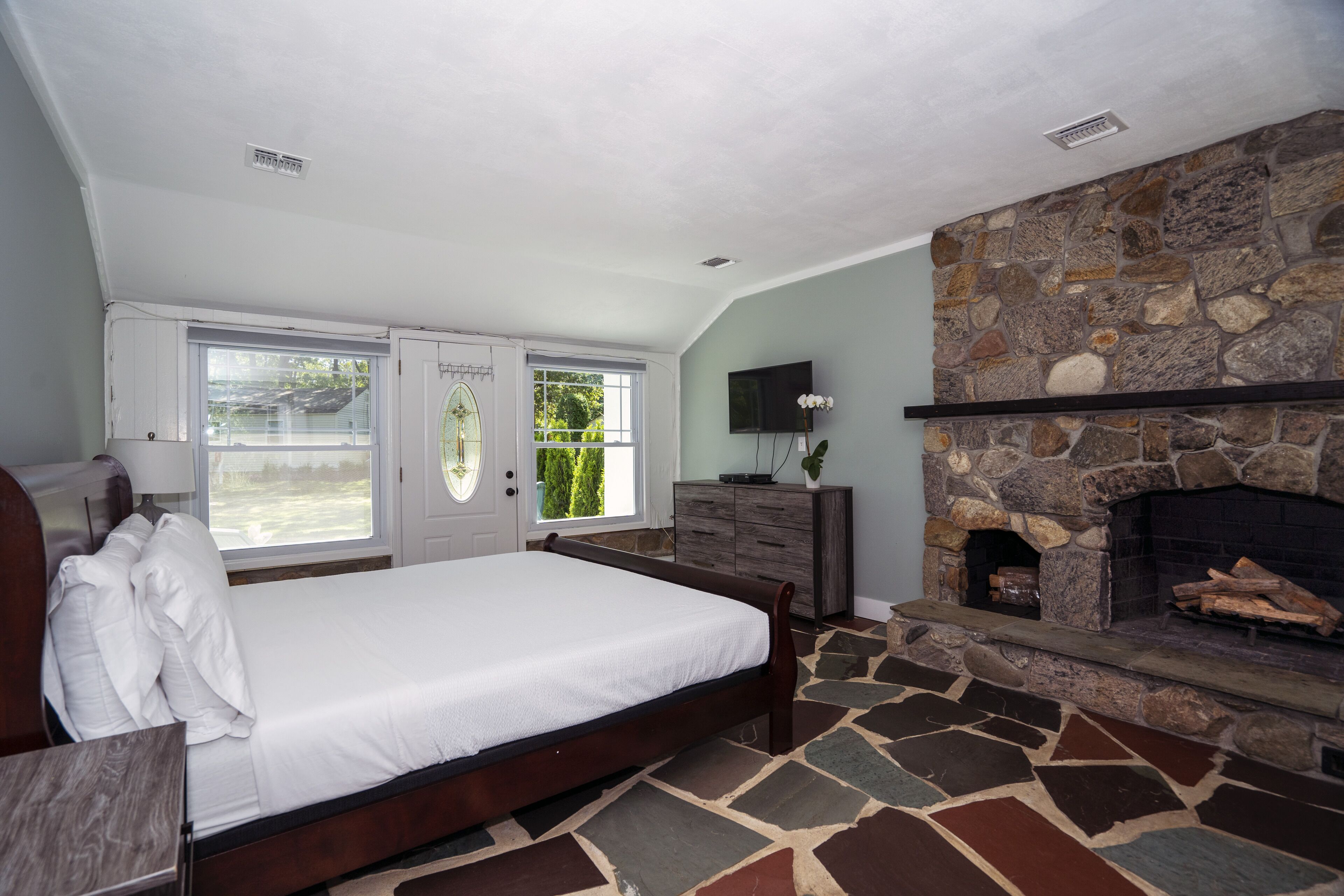 A bedroom with a stone fireplace, a bed with white linens, a dresser, and a TV mounted on the wall.
