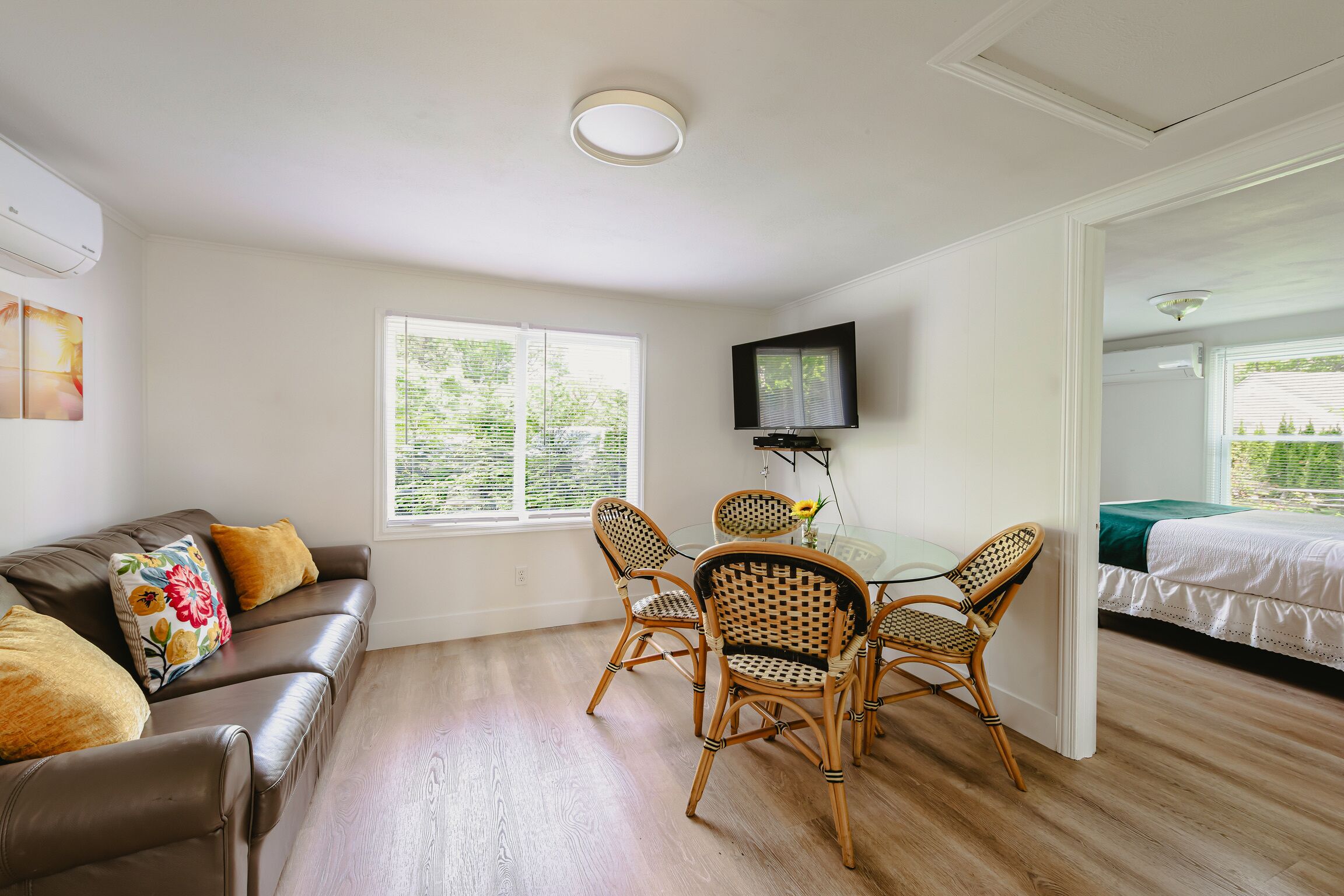 Una habitación con un sofá de cuero marrón, sillas de mimbre, una mesa de vidrio, un televisor montado en la pared y una cama con una manta verde.