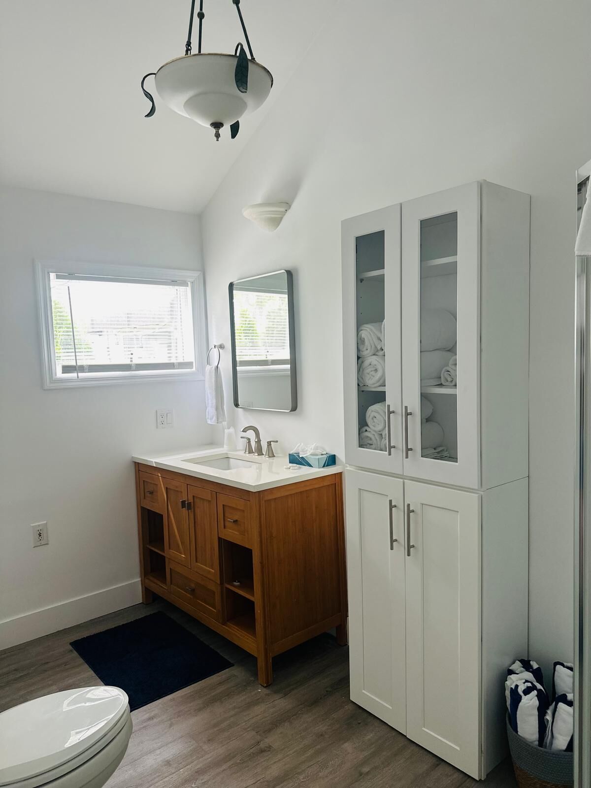 A bathroom with a wooden vanity, a white cabinet with glass doors, a mirror, and a window.