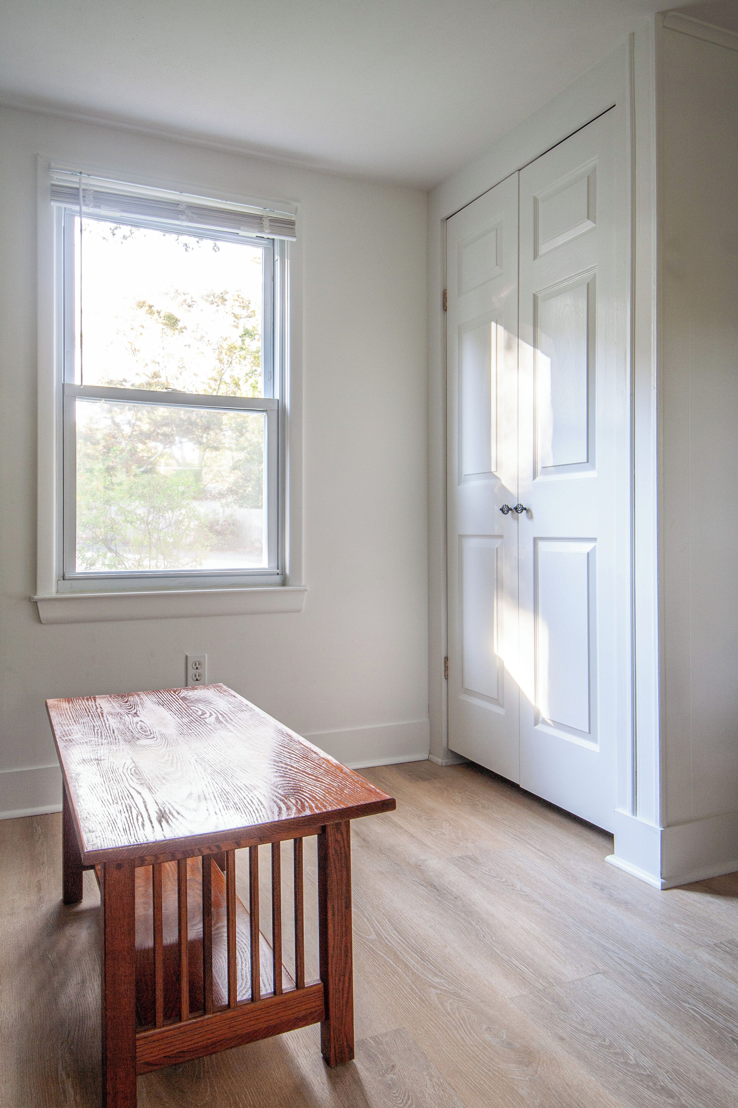 Una habitación con una mesa de madera, una puerta blanca y una ventana con persianas.