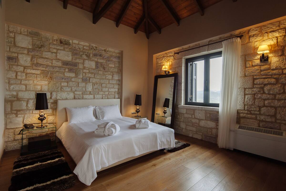 A bedroom with a stone wall, a bed with white linens, two bedside lamps, a large mirror, and a window with curtains.
