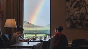 Ein Zimmer mit großem Fenster und Blick auf eine Landschaft, einem Wandbild und einem Tisch mit Stühlen.