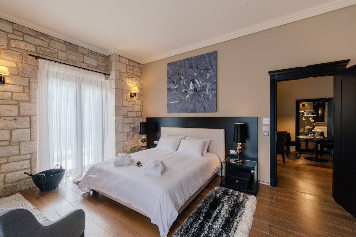 A modern bedroom with a stone wall, a large bed, a painting, and a dining area visible through an open door.