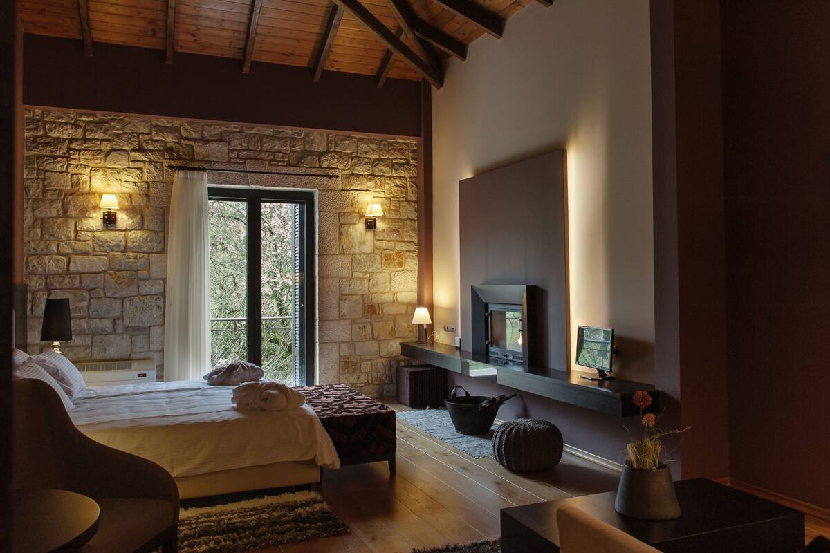 A cozy bedroom with a stone wall, a bed with white linens, a fireplace, and a TV mounted above it.