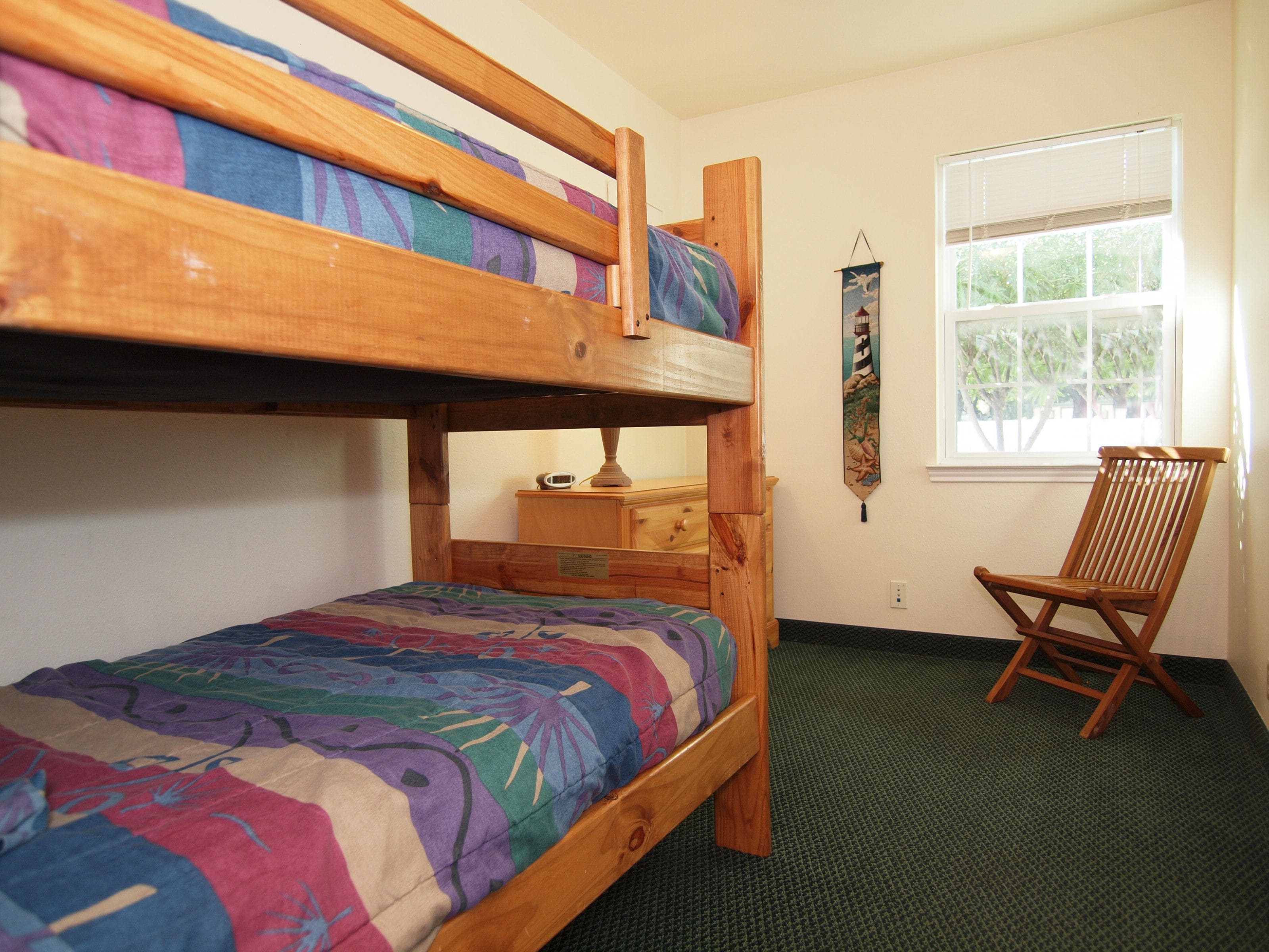A bunk bed room with a green carpet, a wooden chair, and a window with blinds.