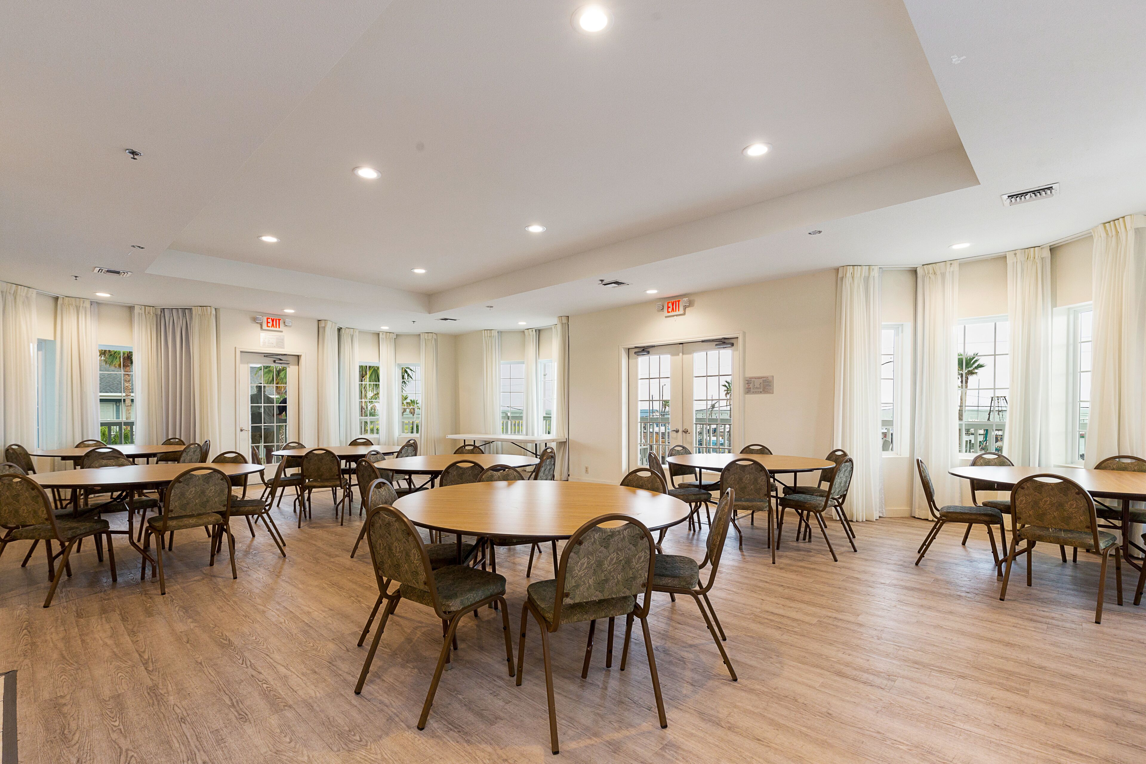 A spacious conference room with multiple tables and chairs, large windows with curtains, and an exit sign on the ceiling.