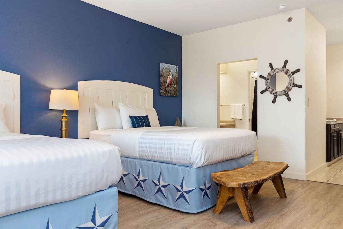 A hotel room with two beds, a wooden bench, a star-patterned bed skirt, a wall-mounted ship's wheel, and a framed picture.