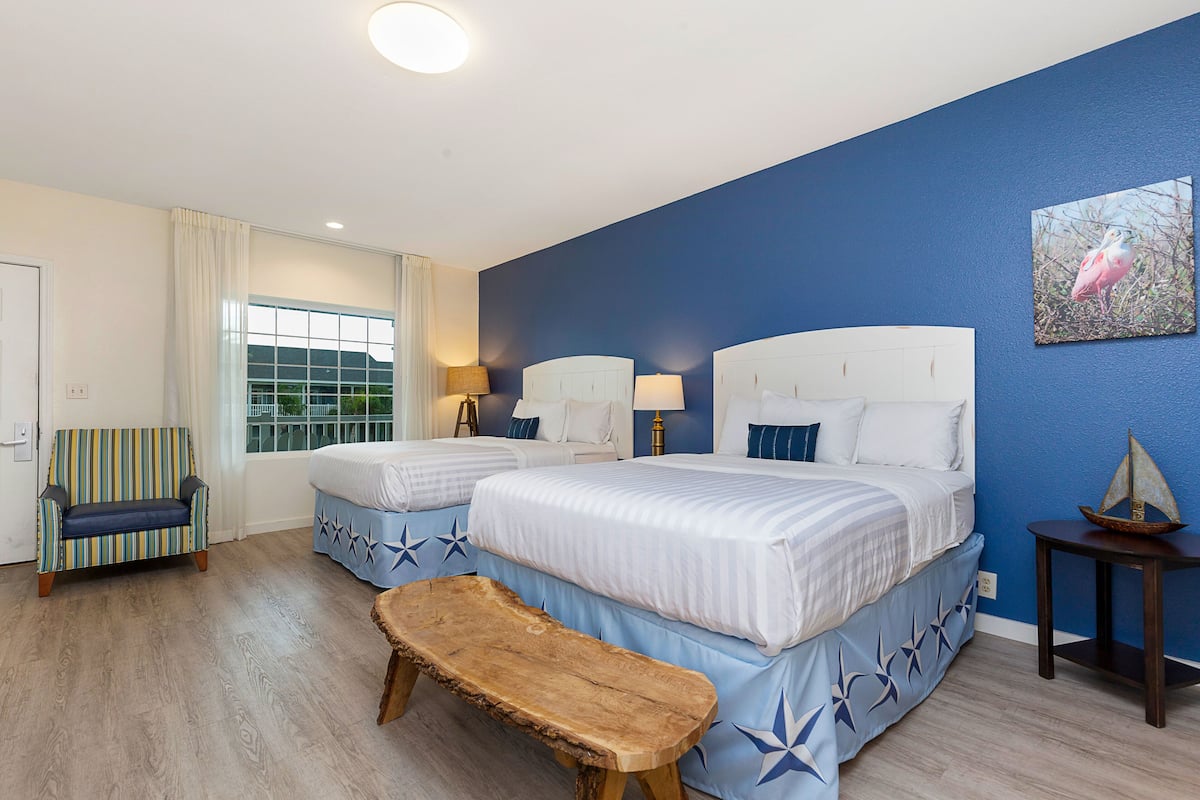 A hotel room with two beds, a bench, a small table, and a window with curtains.