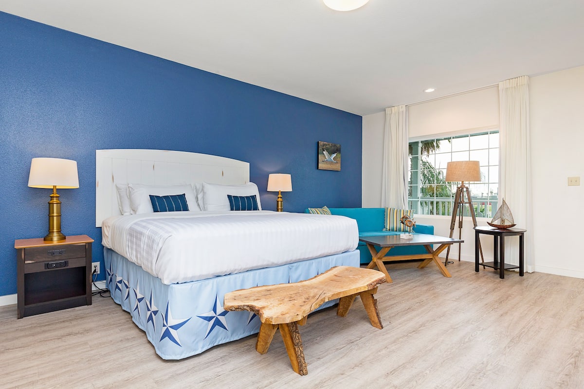 A bedroom with a large bed, two bedside tables with lamps, a wooden bench, a blue sofa, and a small table with a ship model.