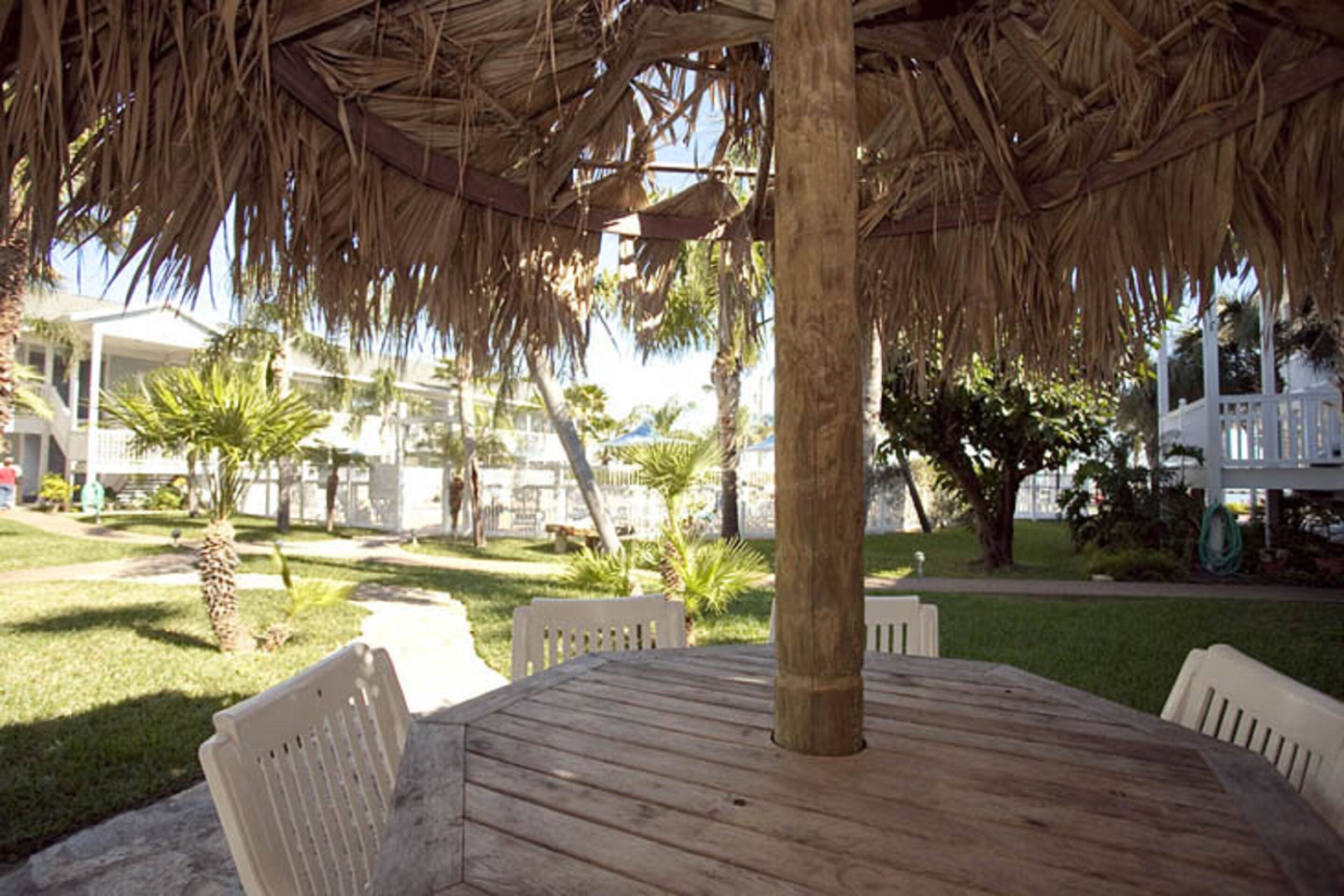 A round wooden table with white chairs under a thatched roof, surrounded by a grassy area with palm trees and a white building in the background.