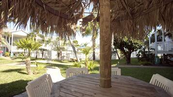 A round wooden table with white chairs under a thatched roof, surrounded by a grassy area with palm trees and a white building in the background.