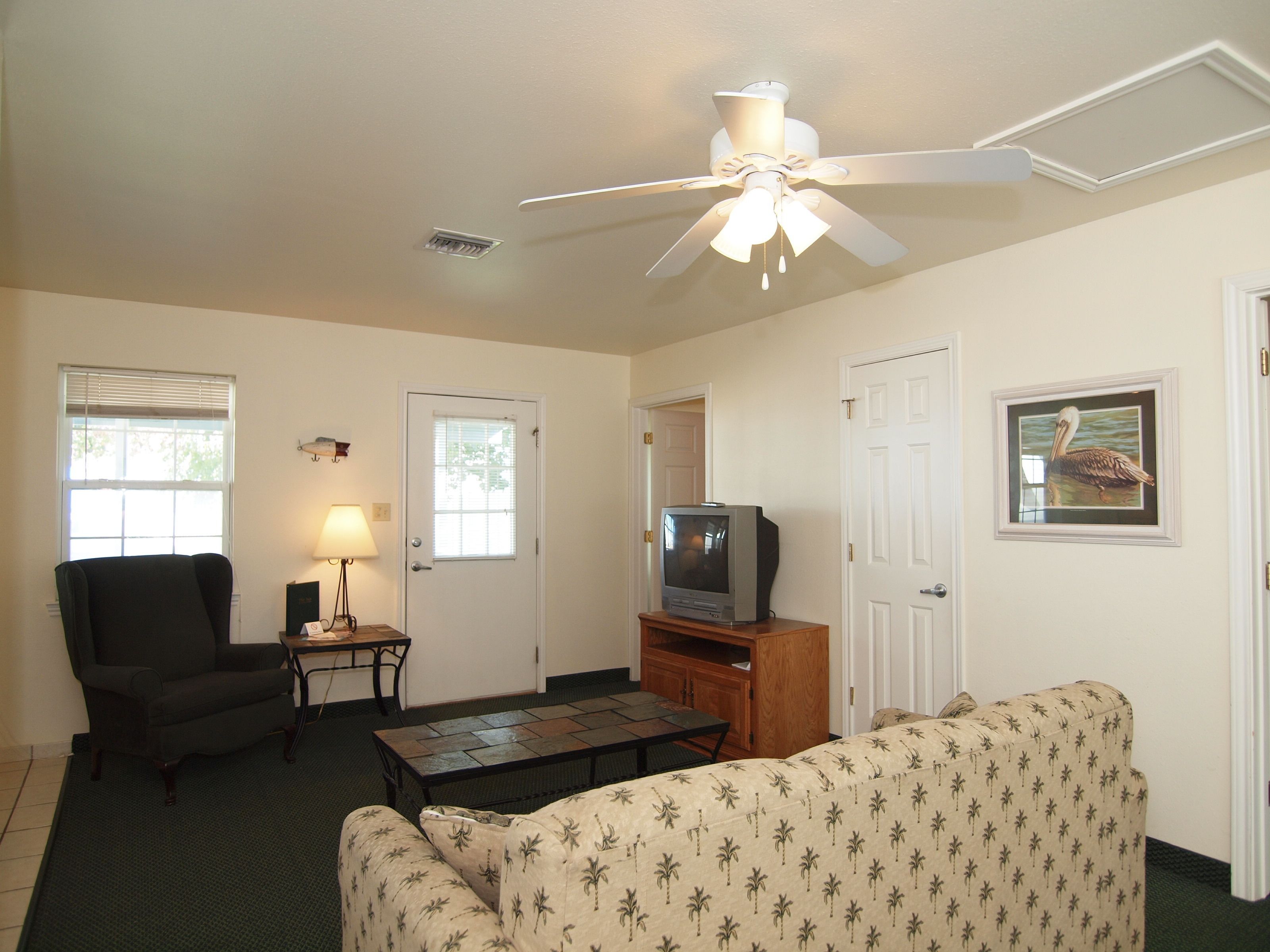 A living room with a ceiling fan, a television on a wooden stand, a black chair, a patterned sofa, and a small table with a lamp.