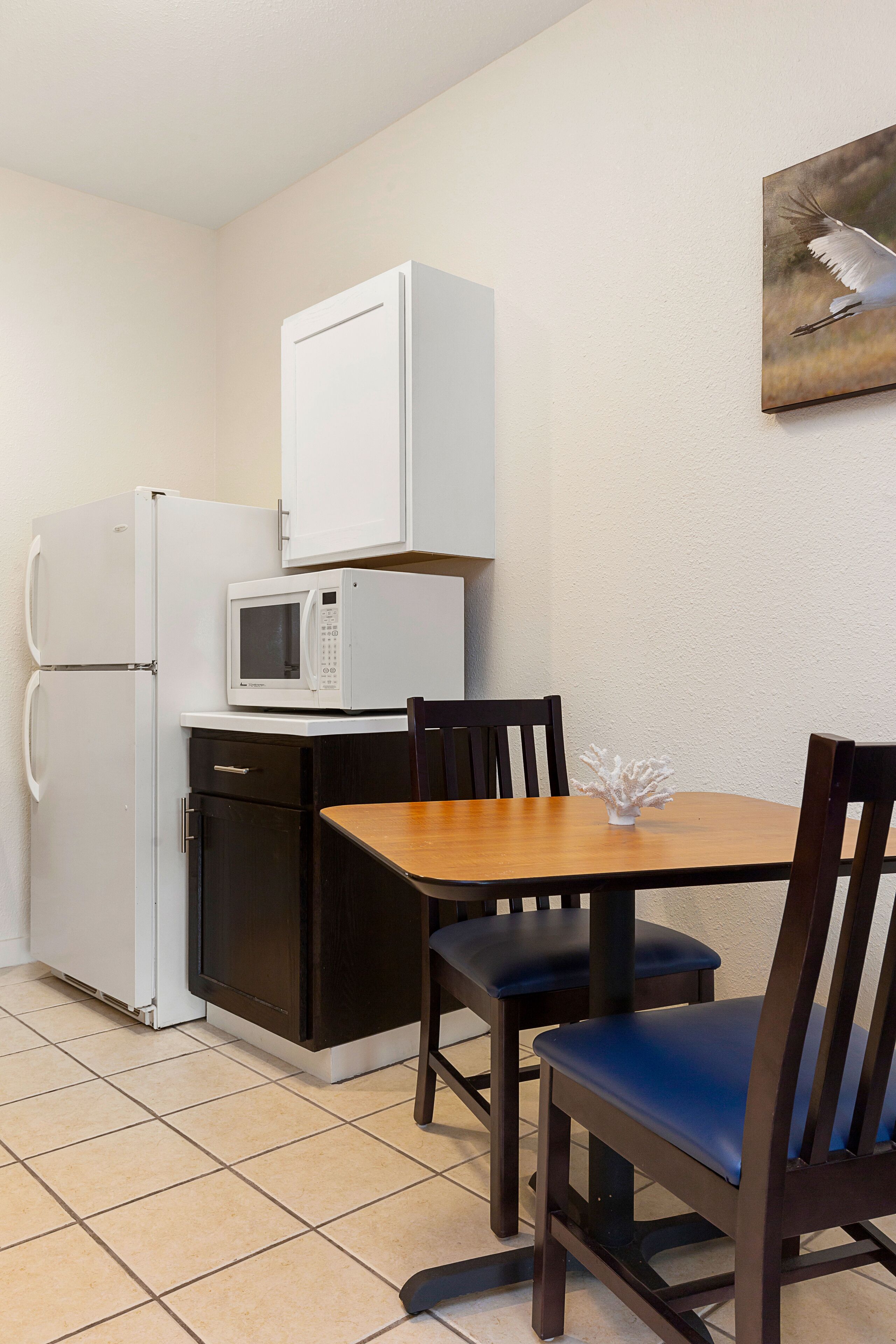 Cocina compacta con refrigerador, microondas y un pequeño armario. Hay una mesa de madera con dos sillas y un florero encima.