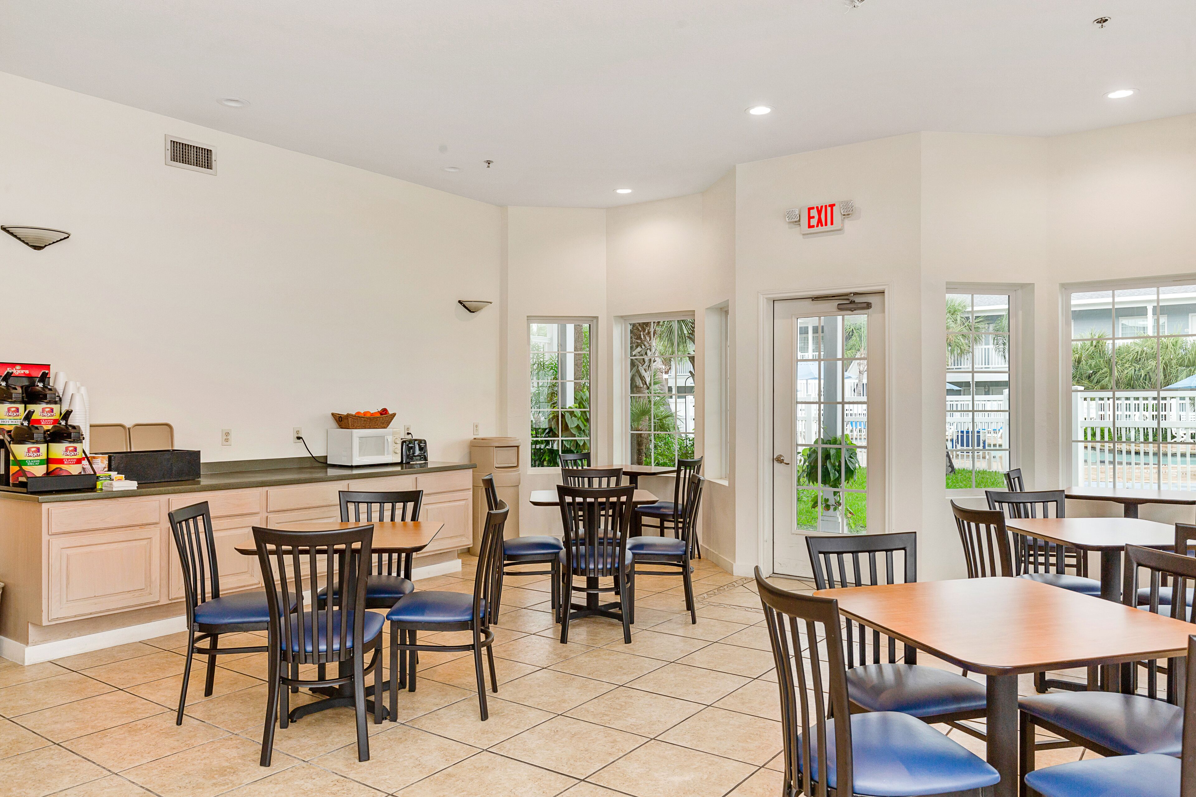 A clean, well-lit dining area with a kitchen counter, several tables and chairs, and large windows offering a view of the outdoors.