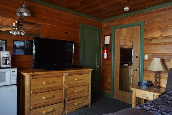 Una habitación con paneles de madera, un aparador de madera, un televisor, un microondas y una mesita de noche con una lámpara.