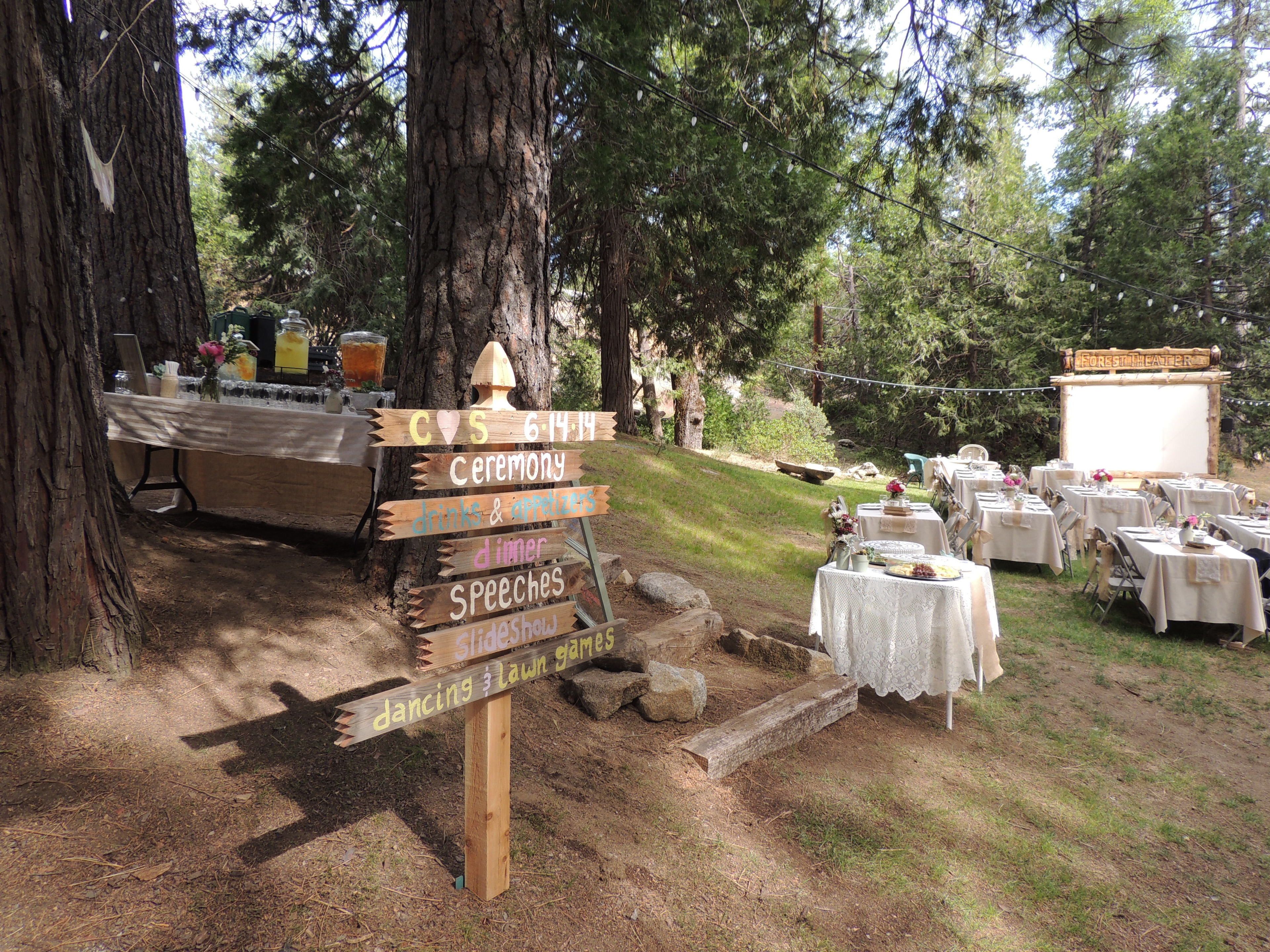 Une décoration rustique pour un mariage en plein air, avec un panneau en bois indiquant les activités prévues, des tables recouvertes de nappes blanches et un décor de fond portant l’inscription « Théâtre de la forêt ».