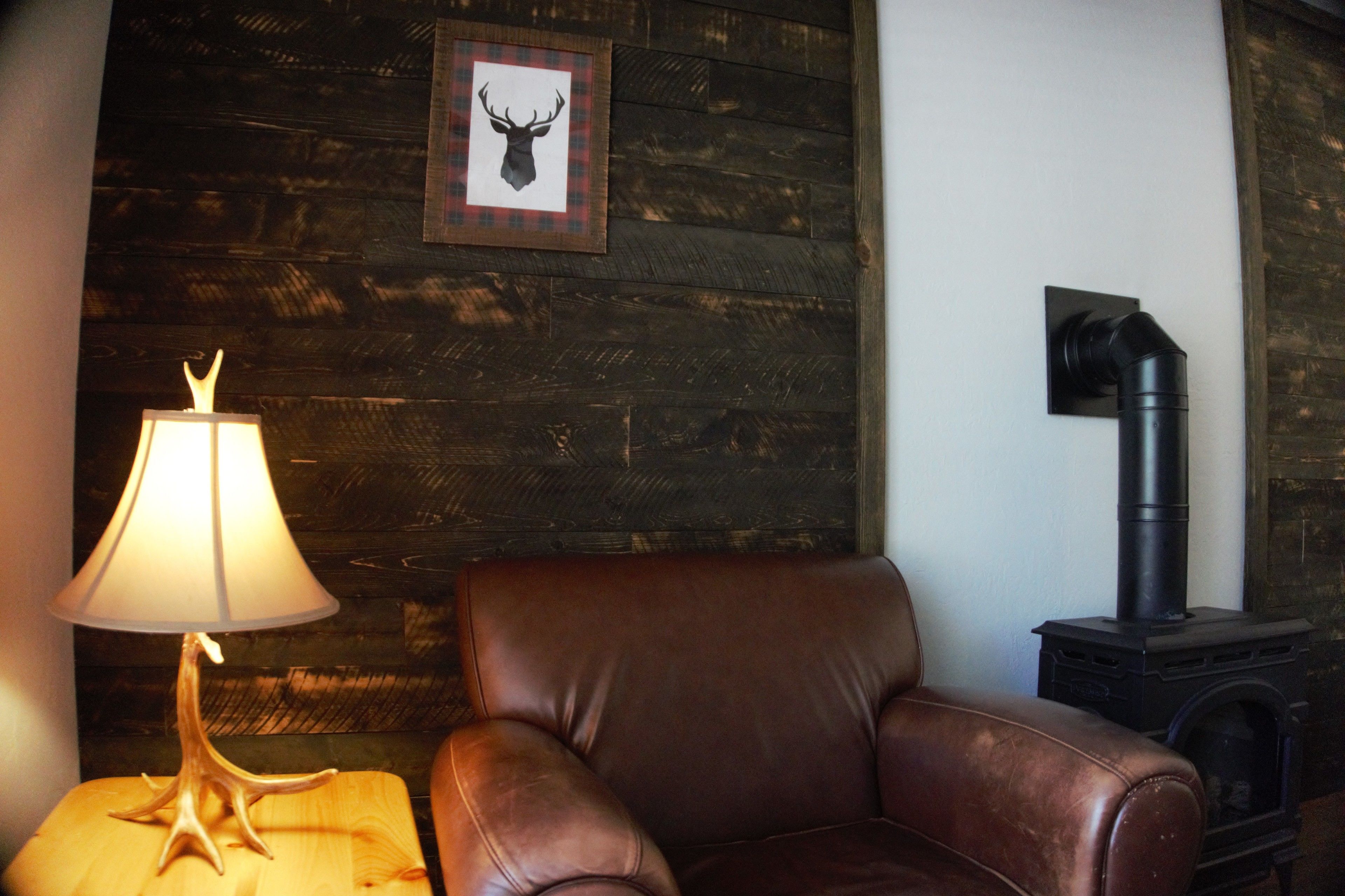 A room with a wooden wall, a framed deer head picture, a brown leather chair, a lamp on a wooden side table, and a black stove.
