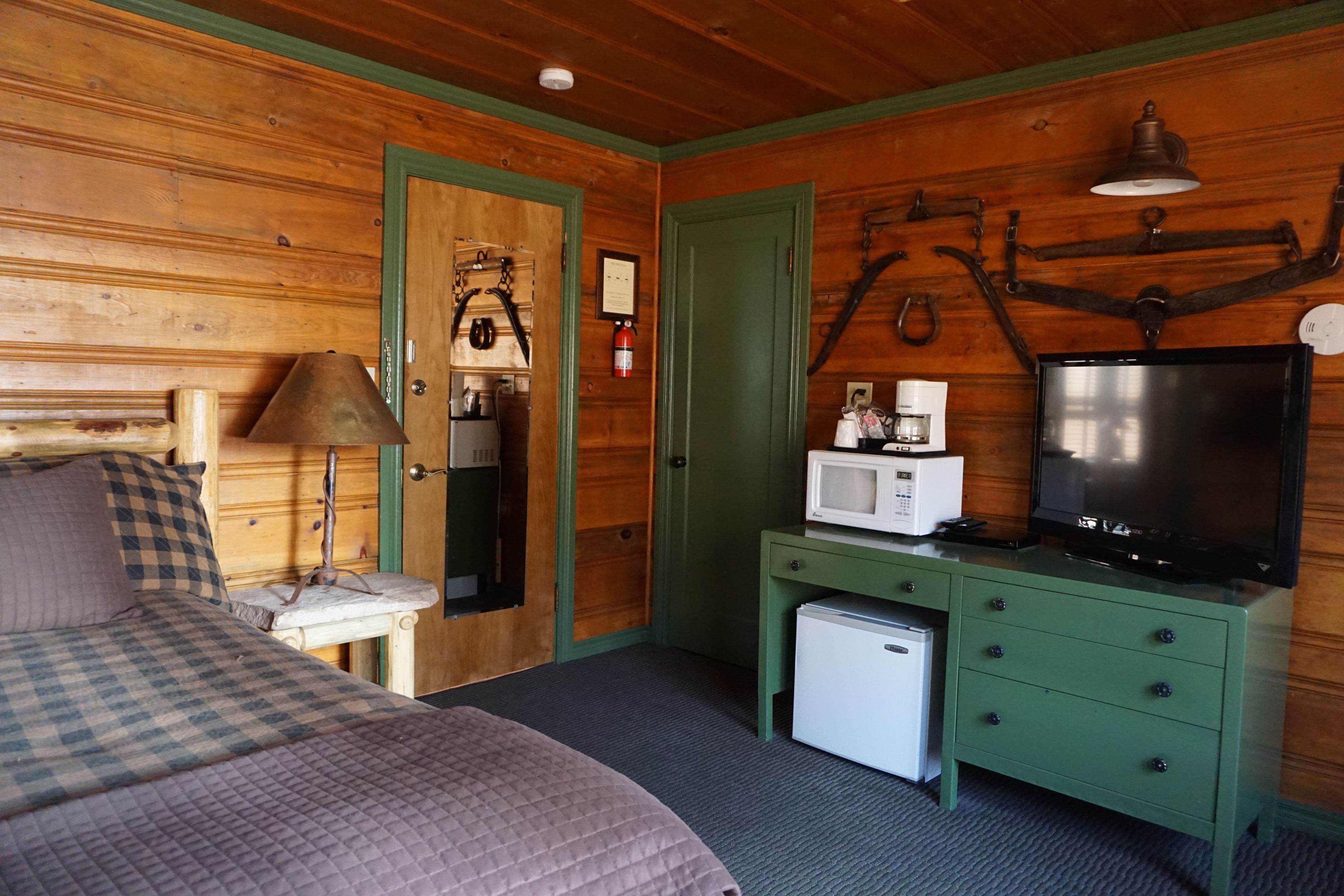 A wooden cabin room with a bed, a desk with a microwave, a television, and a small refrigerator.