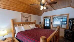Un lit en bois avec un couvre-lit à carreaux rouges, un ventilateur de plafond, une décoration murale représentant un cerf et une fenêtre avec des stores.