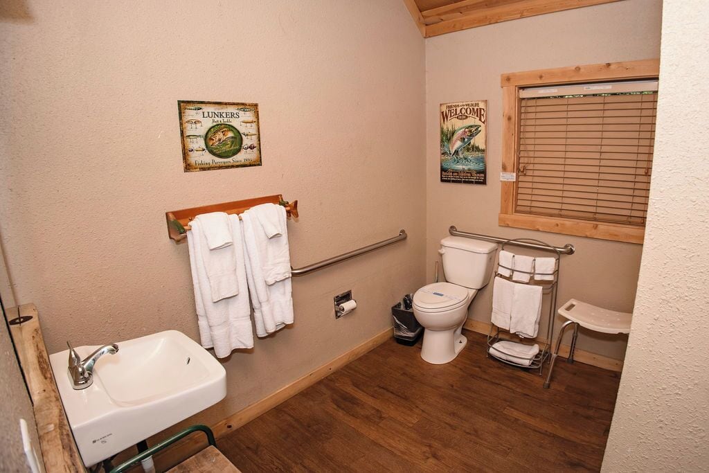 Une petite salle de bain avec un lavabo, des toilettes et une fenêtre avec des stores. Des serviettes sont suspendues sur un porte-serviettes et il y a un panneau accroché au mur.