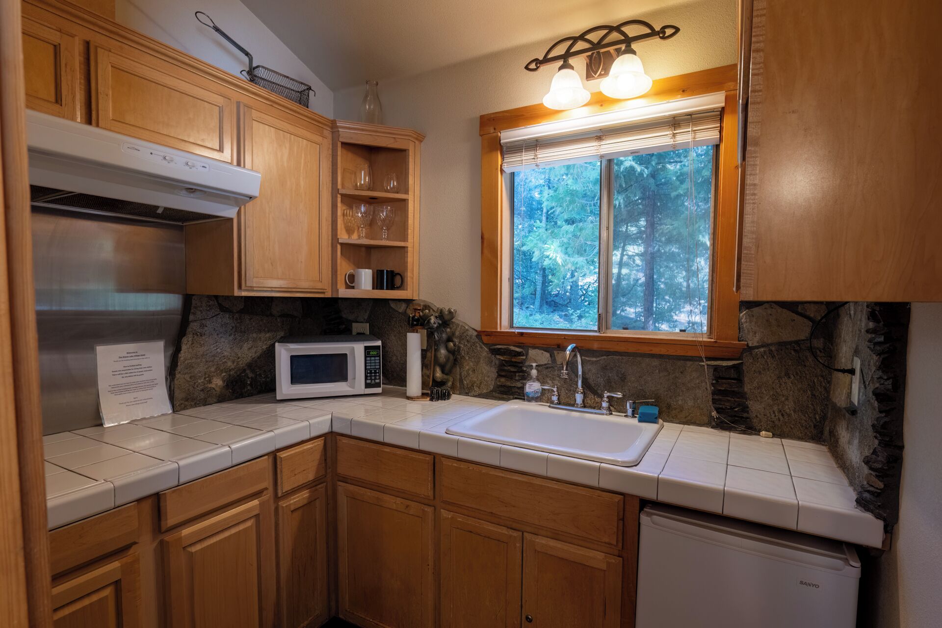 A kitchen with wooden cabinets, a white microwave, a sink, and a window with a view of trees.
