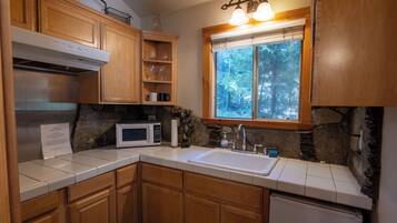 A kitchen with wooden cabinets, a white microwave, a sink, and a window with a view of trees.