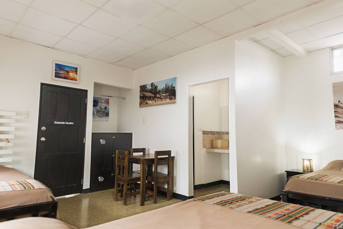 A hotel room with a bed, a desk, a chair, a small table, and a wall-mounted shelf.