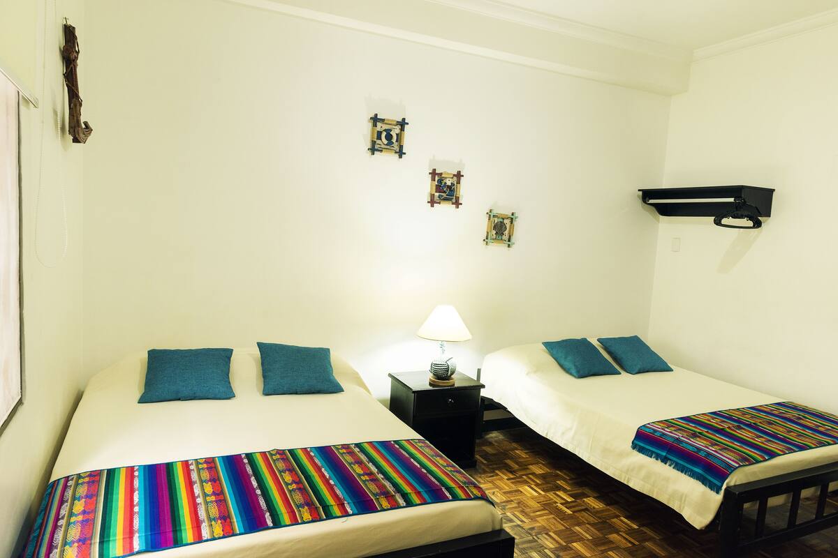 A room with two beds, each with a colorful blanket and blue pillows, a small bedside table with a lamp, and a wall-mounted shelf.