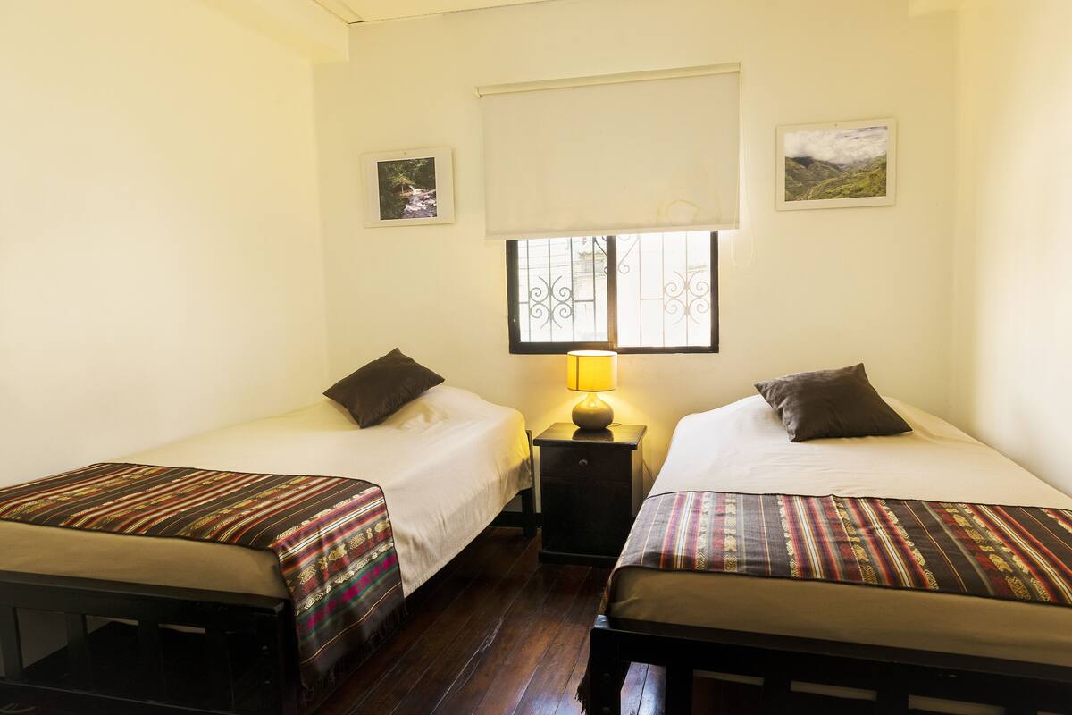 A room with two single beds, a small table with a lamp, and a window with decorative bars.