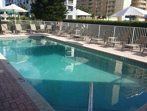 A rectangular swimming pool with a clear blue water surface, surrounded by a white metal fence. There are lounge chairs and umbrellas along the poolside. - Shores Terrace (Pompano Beach)