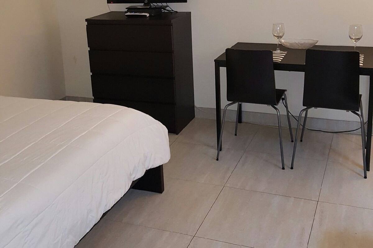 A hotel room with a bed, a TV on a dresser, a desk with two chairs, and a small table with two wine glasses.